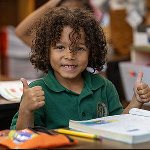 young boy student two thumbs up smiling at camera in class